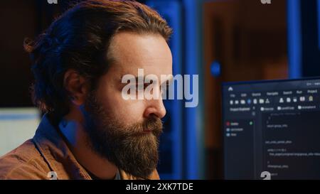 Software technician in apartment office deeply immersed in coding on computer at desk, close up. IT freelancer absorbed by work, focused on screen, writing lines of code at workstation, camera A Stock Photo