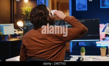 Software engineer writes binary code scripts on computer and drinks coffee. IT specialist in home office enjoying hot beverage while using programming to safeguard consumer data, camera A Stock Photo