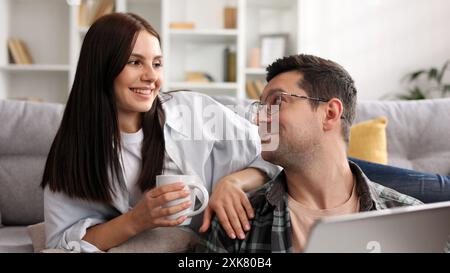 Cheerful young couple relaxing together with laptop and coffee in living room, happy young couple relaxing at home having hot drinks, using computer Stock Photo