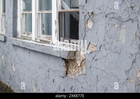 An old grey concrete building with peeling paint and a damaged window sill. The acrylic latex coating is peeling from the wall due to water damage. Stock Photo