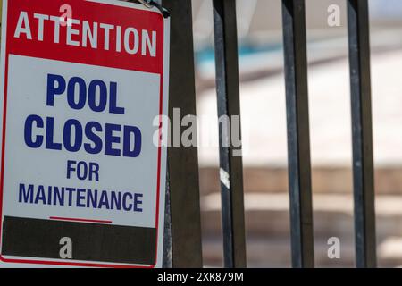 Attention pool closed for maintenance sign posted on a black metal fence with a swimming pool in the background. The pool is closed. Stock Photo