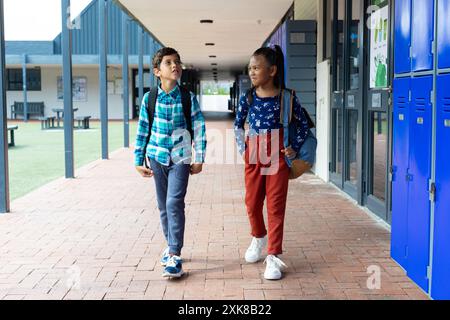 Biracial boy and girl walk past lockers at school. They carry backpacks and engage in conversation, discussing their day. Stock Photo