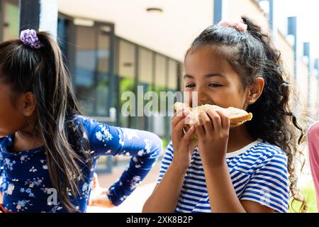 A biracial girl enjoys a sandwich outdoors, eating lunch at school with friends. She is surrounded by peers in a casual, sunny setting, suggesting a b Stock Photo
