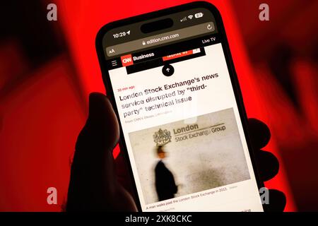 Paris, France - Jul 20, 2024: From a point-of-view perspective, a male hand holds an iPhone displaying a CNN Business article about the London Stock Exchange's news service disruption due to a third-party technical issue, set against a vivid red background Stock Photo