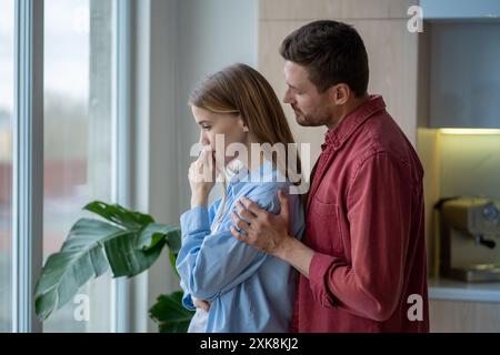 Supportive husband hugging his upset, depressive wife from behind at window in bright apartment Stock Photo