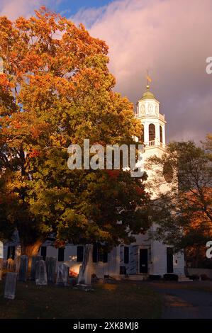 A clearing sky helps illuminate a white steeple  church during autumn. surrounded by fall foliage Stock Photo