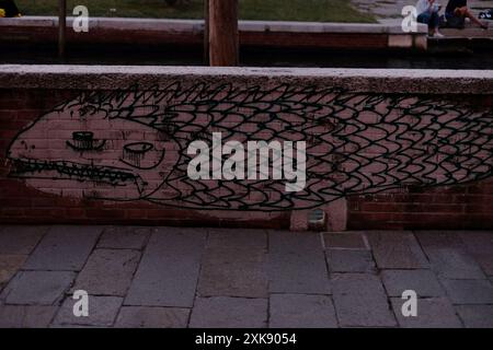 Graffiti of a long, scaled fish, street art on a low brick canal wall of Fondamenta Nani on Dorsoduro, two people sit on the other side,Venice, Italy Stock Photo