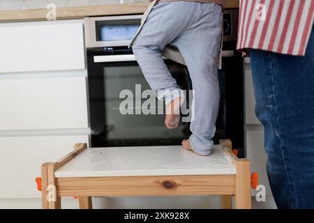 Boy learning how to make pancakes, standing on chair by stove. Parents and children in kitchen, preparing pancake batter, spending weekend day indoors Stock Photo