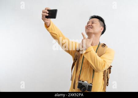 Smiling attractive young tourist with backpack walking and taking selfie mobile phone on white background. Passenger traveling on weekends. Air flight Stock Photo