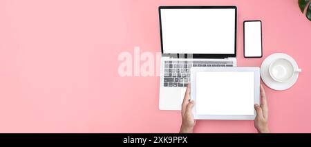 Pastel pink office desk top view cup of coffee, woman hands using laptop, smartphone and holding tablet screen, blank space for advertising, panoramic Stock Photo