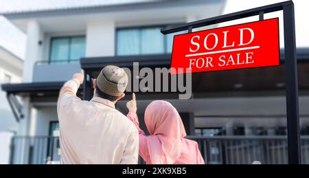 Sold For Sale Real Estate Sign In Front of Property. Portrait of an excited Muslim couple standing outside their new home, walking on the model new ho Stock Photo