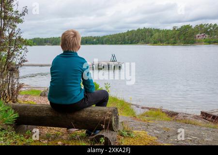 10-years boy on summer vacations having a rest sitting on a wooden log bench near the sea, looking forward the water and forest on the background in a Stock Photo