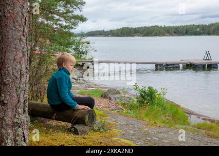 10-years boy on summer vacations having a rest sitting on a wooden log bench near the sea, looking forward the water and forest on the background in a Stock Photo