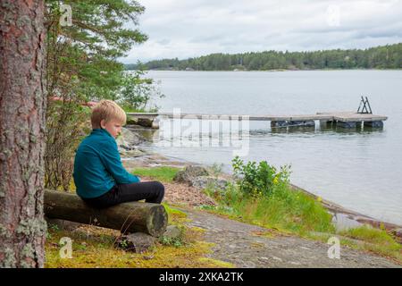 10-years boy on summer vacations having a rest sitting on a wooden log bench near the sea, looking forward the water and forest on the background in a Stock Photo