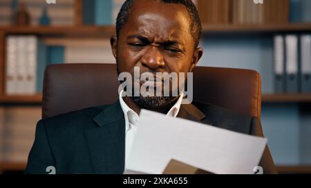 Serious african american businessman sitting at home office opening paper letter upset disappointed man read bad news receives notification with Stock Photo