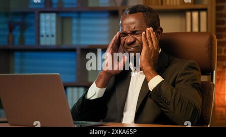 Adult african american businessman working on laptop in office unhealthy tired man suffering from headache feels pain pressure fatigue from overwork Stock Photo