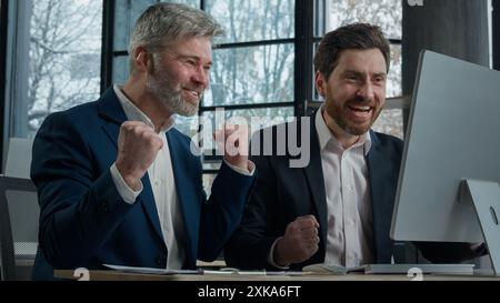 Two businessmen men business partner colleagues collaboration in office working on computer brainstorm in corporate teamwork get good news win deal Stock Photo