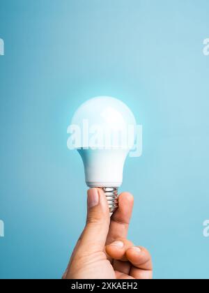 A White LED light bulb in hand, holding isolated on blue background, vertical style. Human finger hold environmentally friendly glowing lamp lightbulb Stock Photo