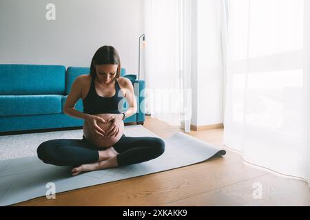Pregnant calm beautiful woman in lotus pose meditating on floor, putting hands heart shape on belly. Motherhood and Wellness Pregnant Concept. Stock Photo