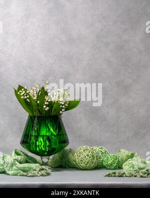 White flowers, lilies of the valley in a green transparent vase, a glass, next to it lies a green scarf and three green balls. Gray background. Plenty Stock Photo