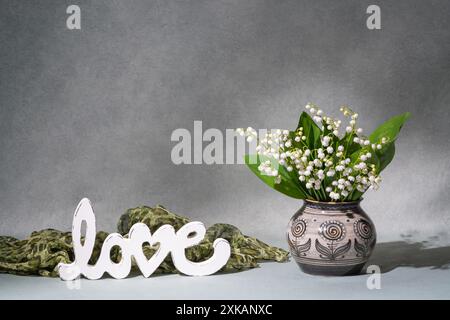 White flowers, lilies of the valley in a vase next to the inscription “love”. Gray background. Plenty of space for text Stock Photo