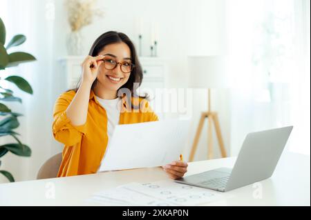 Attractive confident arabian or indian brunette woman, wearing eyeglasses, secretary or financial manager, sits at a table with a laptop, holding documents in hands, looks at camera and smiles Stock Photo