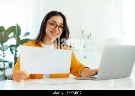 Working process. Positive arabian or indian brunette woman, wearing eyeglasses, secretary or financial manager, sits at a desk with laptop, holding documents in hands, looksa and smiles at camera Stock Photo