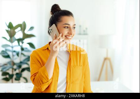 Positive pretty young woman of indian or arabian ethnicity, manager, team leader, seo, talking by mobile phone with client while standing in a modern office space, looking away and smiling Stock Photo