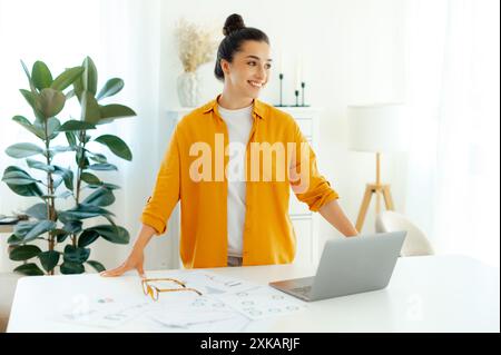 Successful confident pretty brunette arabian or indian ethnicity young woman stylishly dressed, company seo, creative manager, secretary stands near work desk, looks away and smile friendly Stock Photo