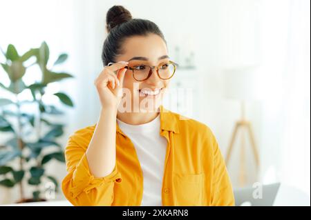 Close-up of a confident gorgeous arabian or indian young woman with eyeglasses, manager, freelancer or student standing indoors near work desk with arms crossed, looking away and smiling friendly Stock Photo