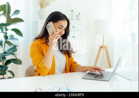 Mobile phone conversation. Positive pretty arabian or indian young woman, product manager, web designer, sits at a desk working in a laptop on a project, having smartphone conversation with a client Stock Photo