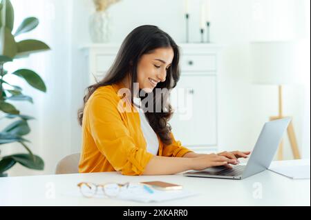 Online work. Positive pretty brunette arabian or indian young woman, IT specialist, product manager, web designer, sits at a workplace working in a laptop on a project, looking at a screen, smiles Stock Photo