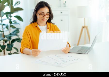 Documentary routine. A beautiful, smart, focused woman of arabian or indian nationality, wearing eyeglasses, sits at a table with a laptop, draws up documents, analyzes financial reports Stock Photo