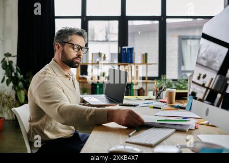 A handsome businessman with a beard works intently at his laptop in a modern office setting. Papers and office supplies surround him on his desk. Stock Photo