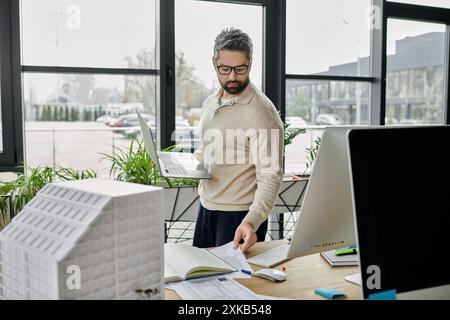 A businessman with a beard works on a laptop and reviews architectural plans in a modern office. Stock Photo