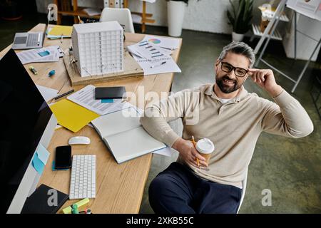 A handsome, bearded businessman in a modern office setting, sits with a coffee in hand and a relaxed smile, taking a break from his work. Stock Photo