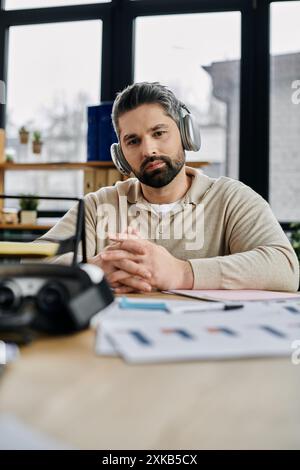 A handsome businessman with a beard sits at a modern office desk, wearing headphones and looking intently at documents. Stock Photo