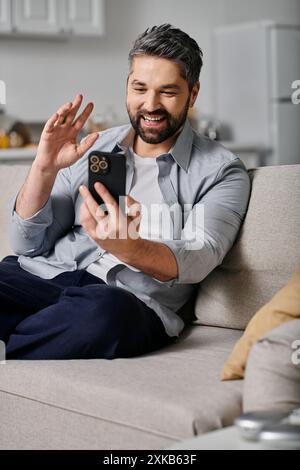 A bearded man in casual attire sits on a couch and waves at his phone while video chatting. Stock Photo