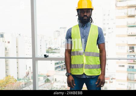 Portrait of young African man engineer standing in office looking at camera, he is wearing reflective helmet and yellow safety helmet, he has window b Stock Photo