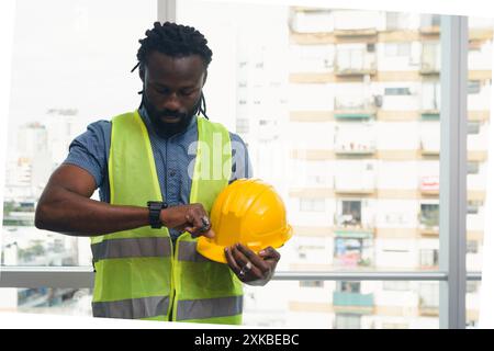 Young African engineer Man with helmet and vest is standing in office in front of window checking time on his watch to leave. Stock Photo