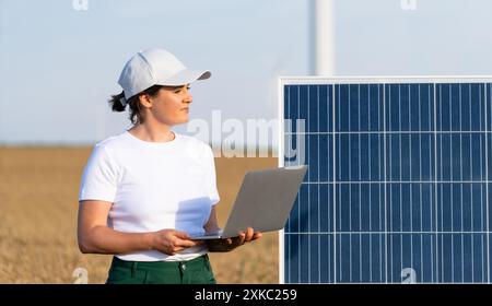 Woman farmer wearing white cap and t-shirt with laptop stands next to solar panel. Wind turbines in the background. Stock Photo