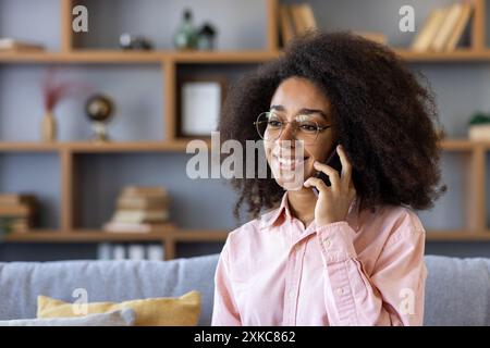 Happy woman with curly hair and glasses talking on smartphone while sitting on a couch. Cozy home setting with wooden shelves and books in the background. Casual conversation, relaxed atmosphere. Stock Photo