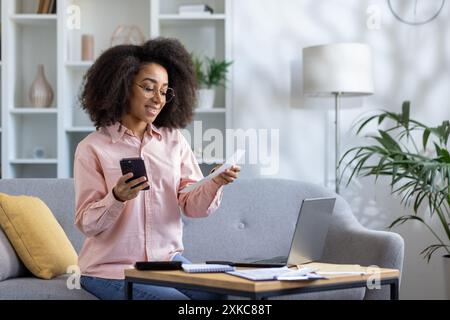 Smiling woman using smartphone and laptop for online payment at home office. Concept of digital banking, remote work, and financial management in modern living room setup. Stock Photo