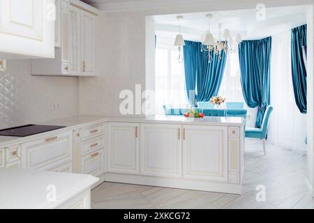 A sophisticated white kitchen with gold accents and a view of an elegant dining area with blue velvet chairs and curtains. The interior design showcas Stock Photo