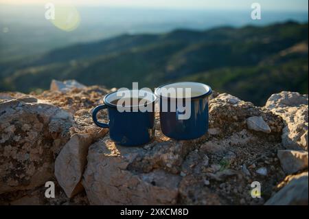 Two blue enamel mugs filled with hot beverages sit on a rocky surface, with a scenic mountain view in the background during sunrise. Stock Photo