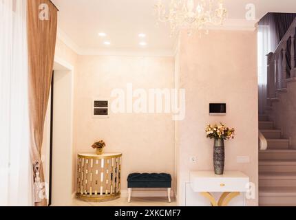 A well-lit corner featuring a gold console table with a flower vase, a turquoise bench, and a white drawer unit with a decorative vase. The chandelier Stock Photo