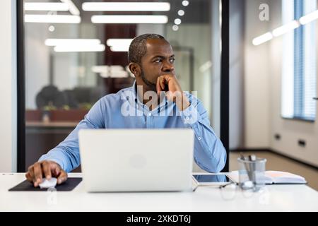 Professional businessman sitting at office desk with laptop, deep in thought. Modern workspace with natural light. Concept of business planning, strategy, and focus. Stock Photo