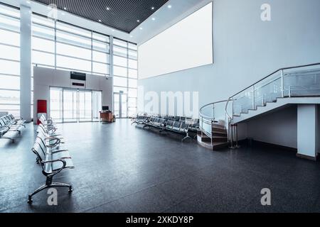 Spacious airport waiting area with rows of empty chairs, large windows providing natural light, a digital clock, and a staircase leading to the upper Stock Photo