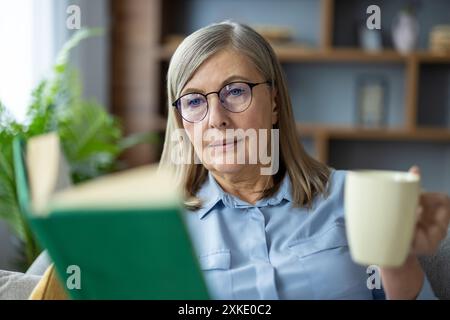 Senior woman wearing glasses reading book while drinking coffee at home. Relaxed elderly lady enjoying quiet time and leisure activity in cozy living room environment. Stock Photo
