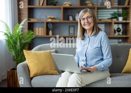 Smiling senior woman working on laptop sitting on sofa. Comfortable home setting with bookshelves and plants in background. Concept of remote work, te Stock Photo